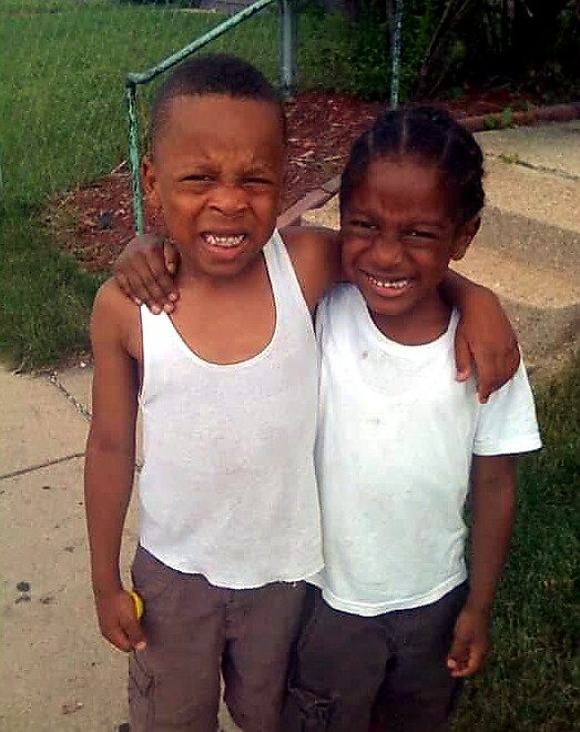 Two smiling boys with arms around each other pose for a photo on a sidewalk near a fence. One wears a white tank top, the other a white tee.