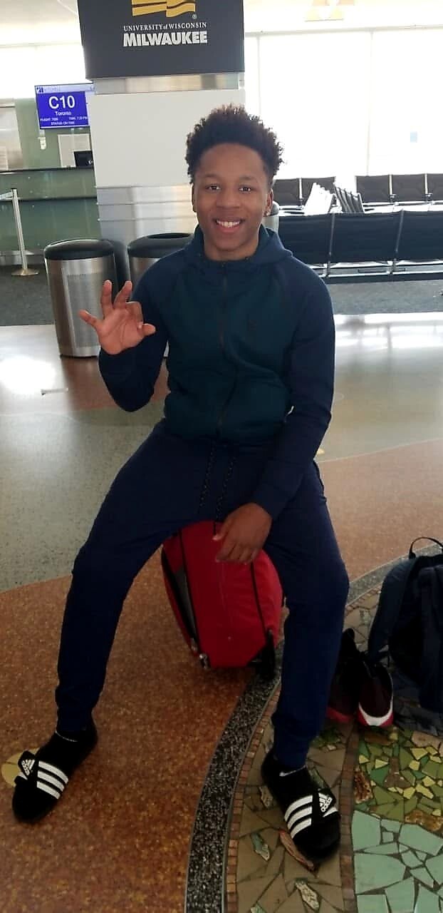 Smiling young man in a navy tracksuit sits on a red backpack, waving in a waiting area with a University of Wisconsin–Milwaukee sign visible.