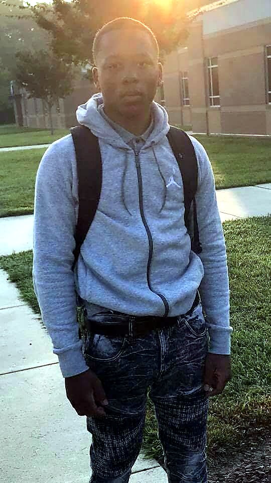 Young man in a gray hoodie with a black backpack standing on a sidewalk near a campus building, sun glare behind him.