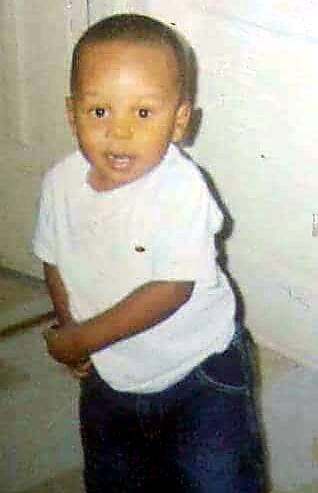 Young boy indoors, standing and smiling, wearing a white T-shirt and dark jeans with his arms bent.