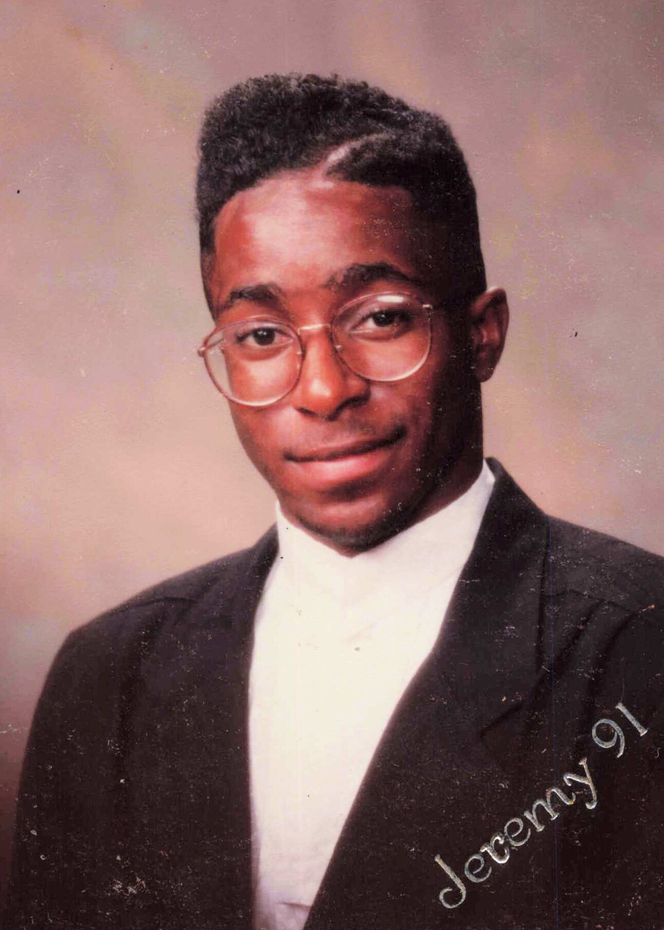 Formal portrait of a young man in a dark suit and white shirt, wearing round glasses and a short, neatly styled afro.