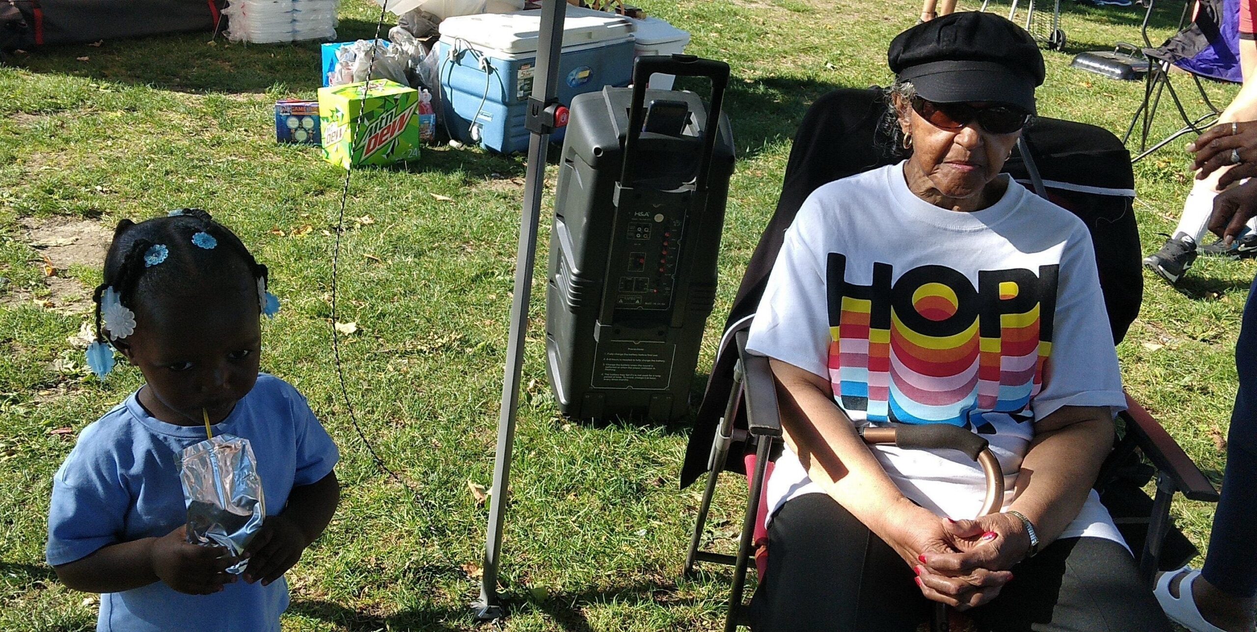 A young girl with blue hair accessories drinks from a foil pouch outdoors while a woman in a wheelchair sits nearby in a park setting with a cooler and speaker behind them.