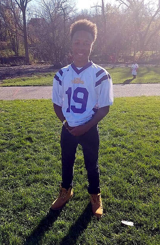 Young person in a white football jersey numbered 19 stands on grass in a park, smiling at the camera with sunlight behind.