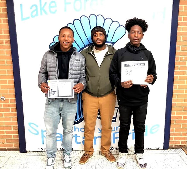 Three men stand side by side in front of a school mural; the two on the ends hold certificates and smile at the camera, with a supervising man in the middle wearing a green jacket.
