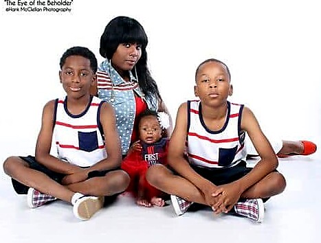 African American mother with two sons in coordinated outfits and an infant, seated on the floor against a white backdrop.