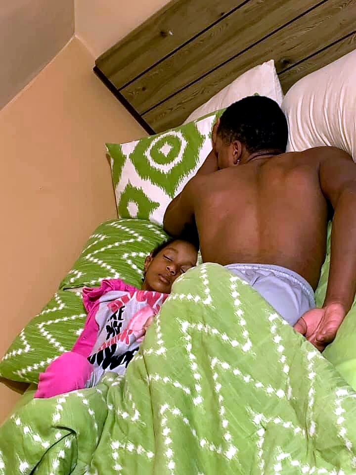 An adult man sits at the edge of a bed, shirtless, next to a child sleeping under a green patterned blanket.