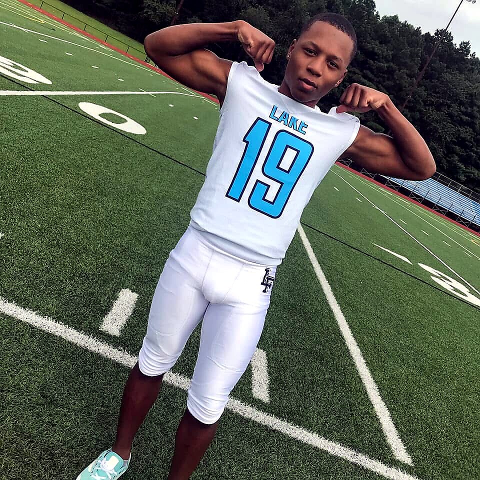 Youth football player on a turf field, wearing a white jersey with 'LAKE' and number 19, flexing his biceps.