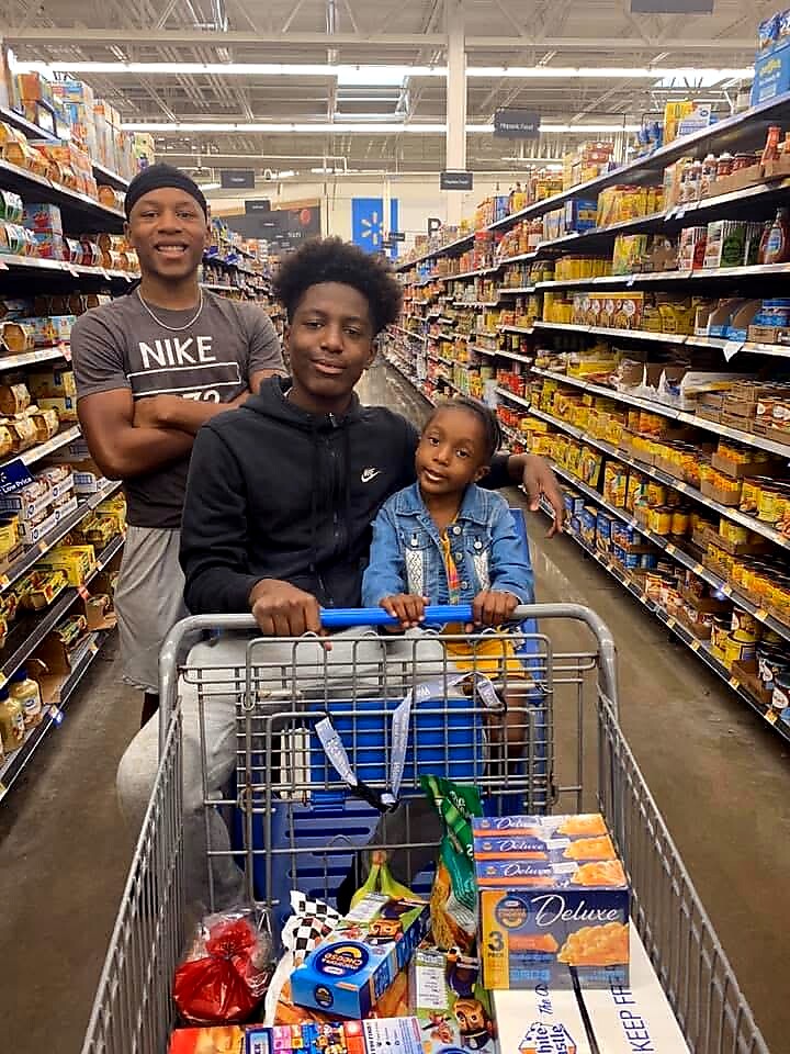 Three siblings posing and smiling in a grocery store aisle with a cart full of groceries behind them.