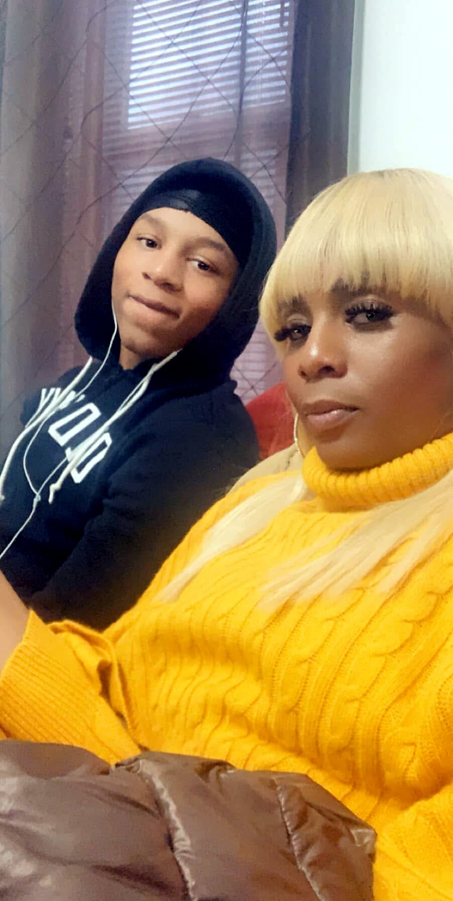Two people posing for a selfie indoors: a woman in a bright yellow knit sweater in the foreground and a young man in a black hoodie in the background behind her.