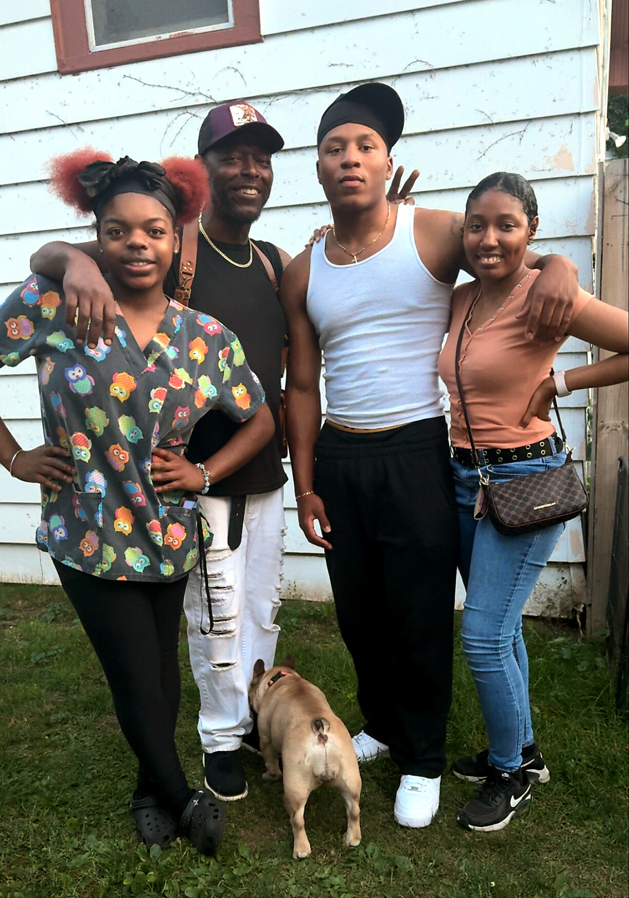 Group of four friends posing closely together outside a light-blue clapboard house; a small tan dog stands at their feet.