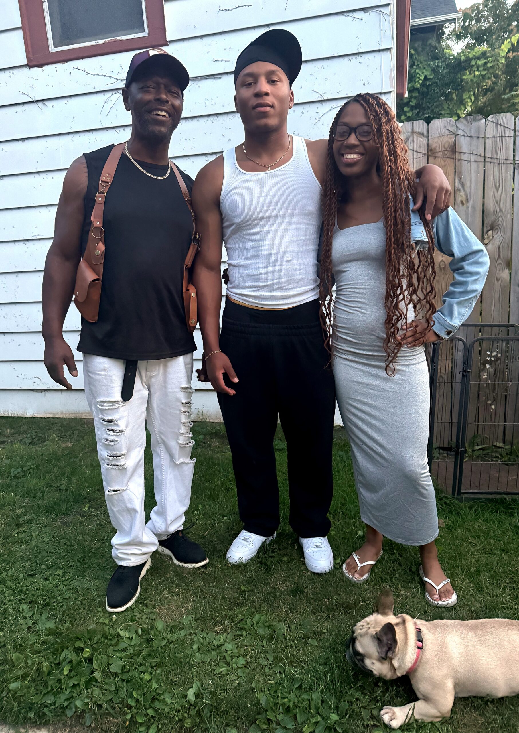 Three friends posing outdoors in front of a light siding house; left man in black sleeveless shirt and white ripped jeans, center person in white tank top and black pants, right woman in gray dress with a denim jacket, a bulldog on the grass in the foreground.