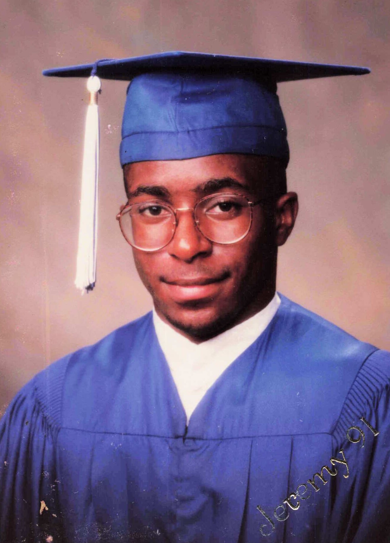 Young man in a blue graduation cap and gown with round glasses, facing the camera.