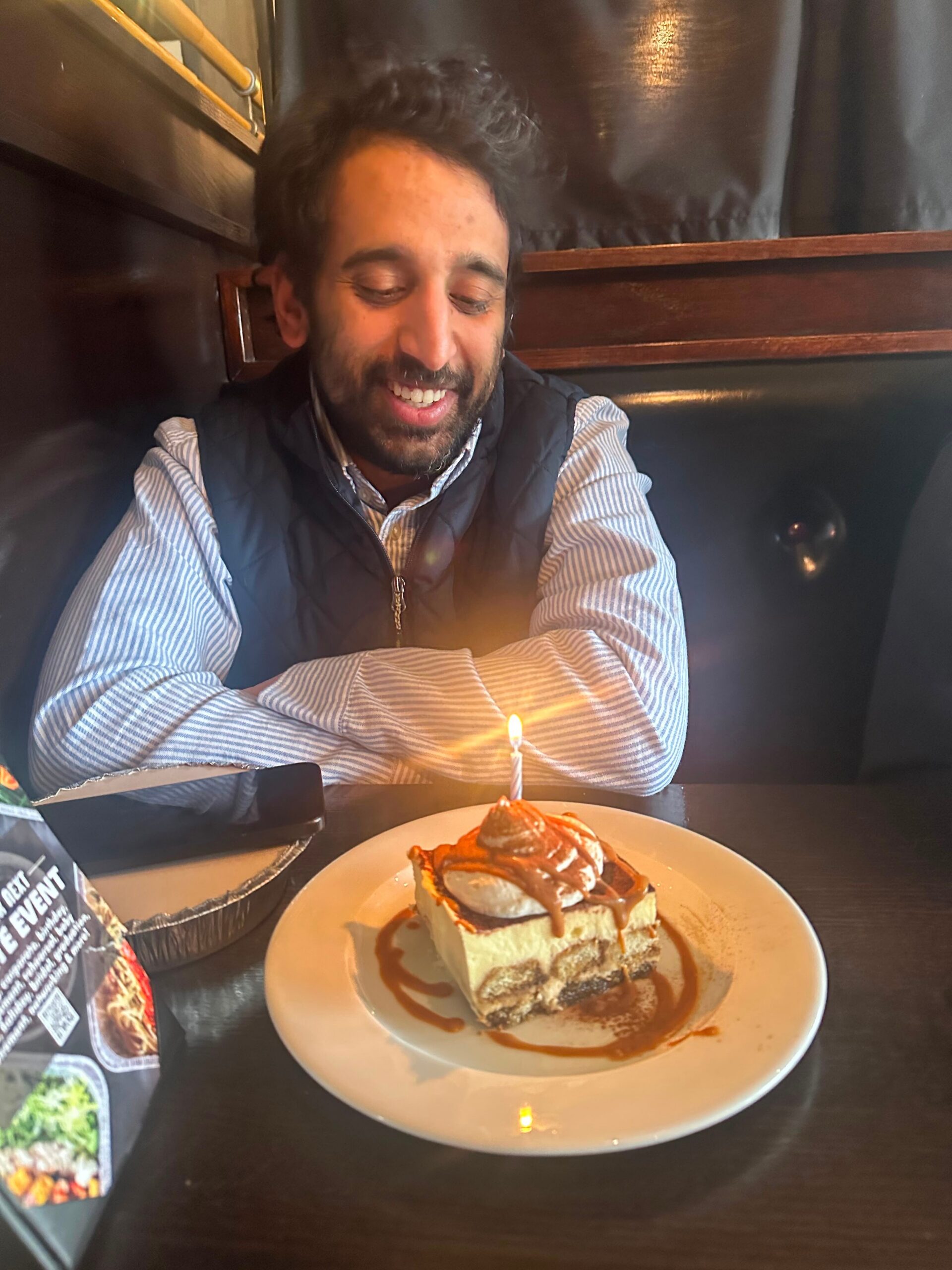 Man smiling at a birthday cake with a lit candle, seated at a dark wooden table in a restaurant; wearing a striped shirt and dark vest.
