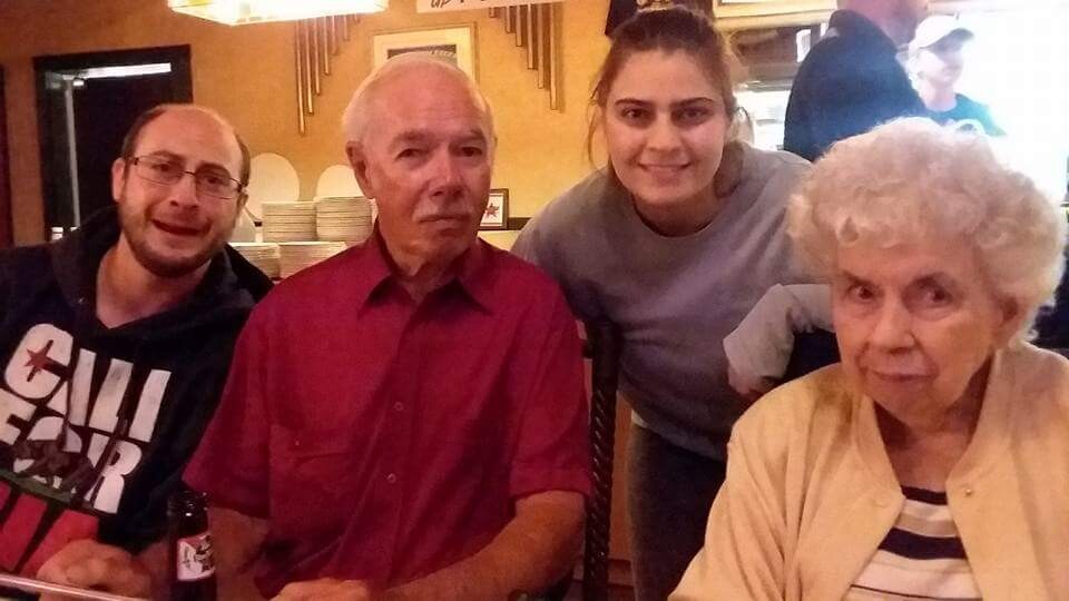 Group photo of four people at a restaurant table: an older man in a red shirt, a smiling young woman, a man in glasses, and an elderly woman in a beige cardigan.