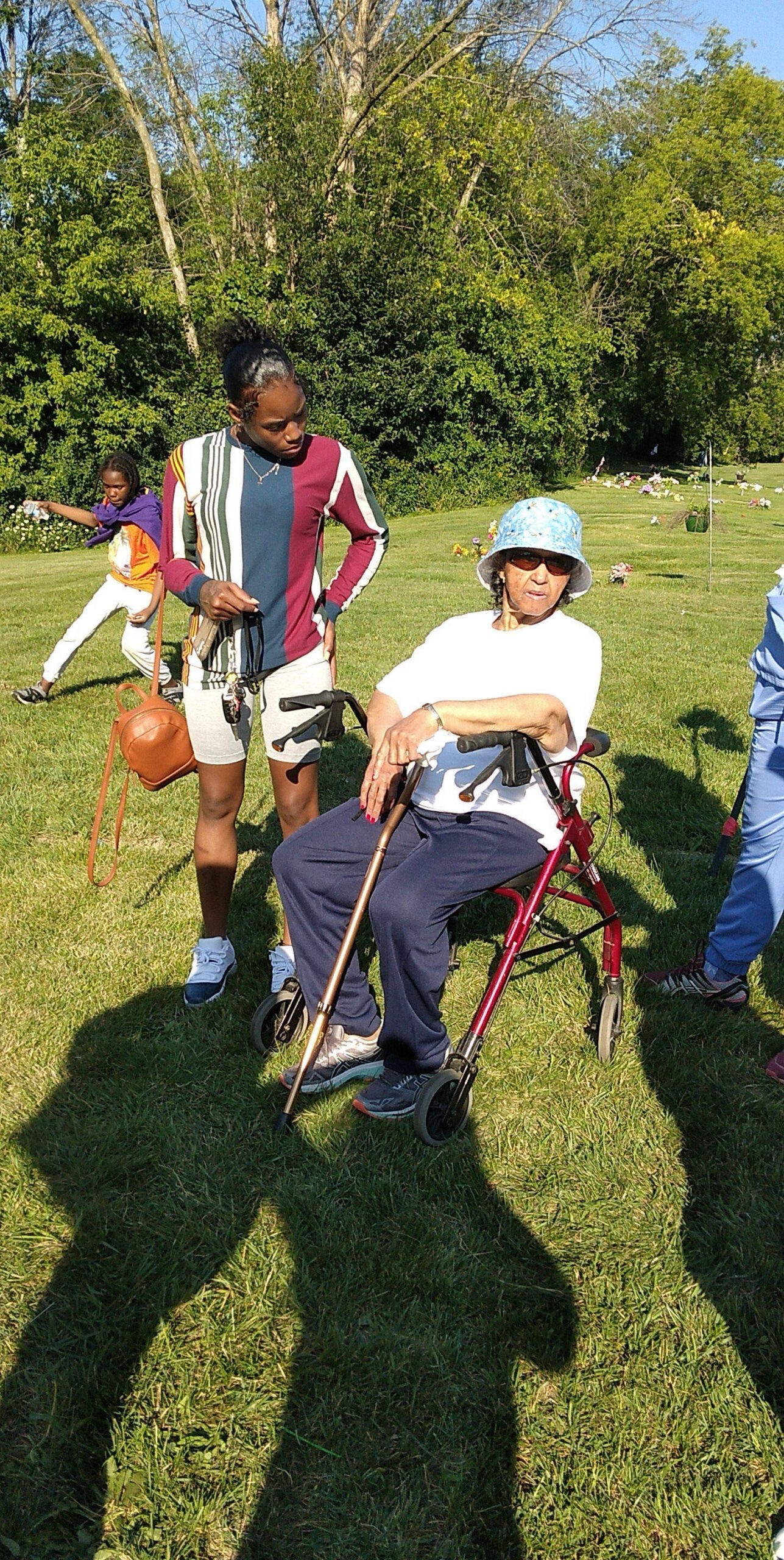 Young girl standing beside an elderly woman in a red wheelchair on a sunny grassy area with trees in the background, likely a cemetery or park setting.