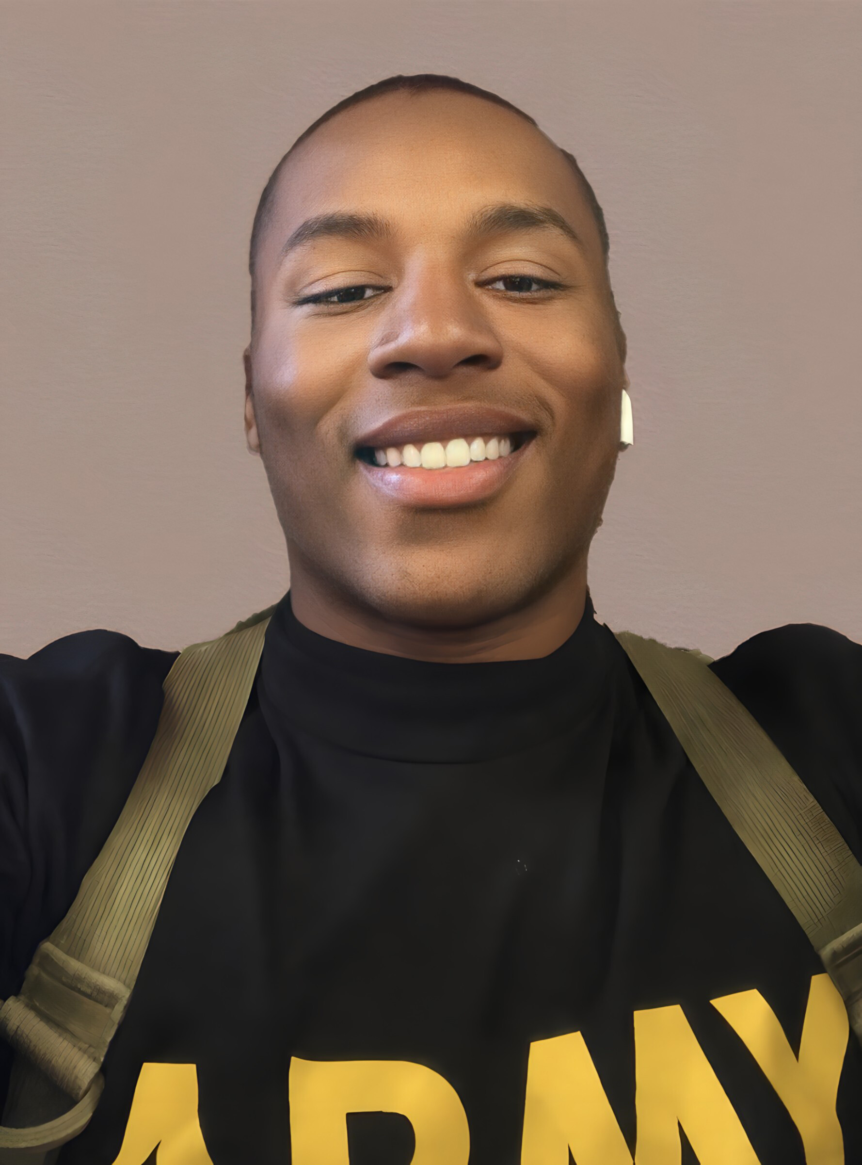 Close-up selfie of a smiling person wearing a black shirt and tan suspenders against a light brown wall.