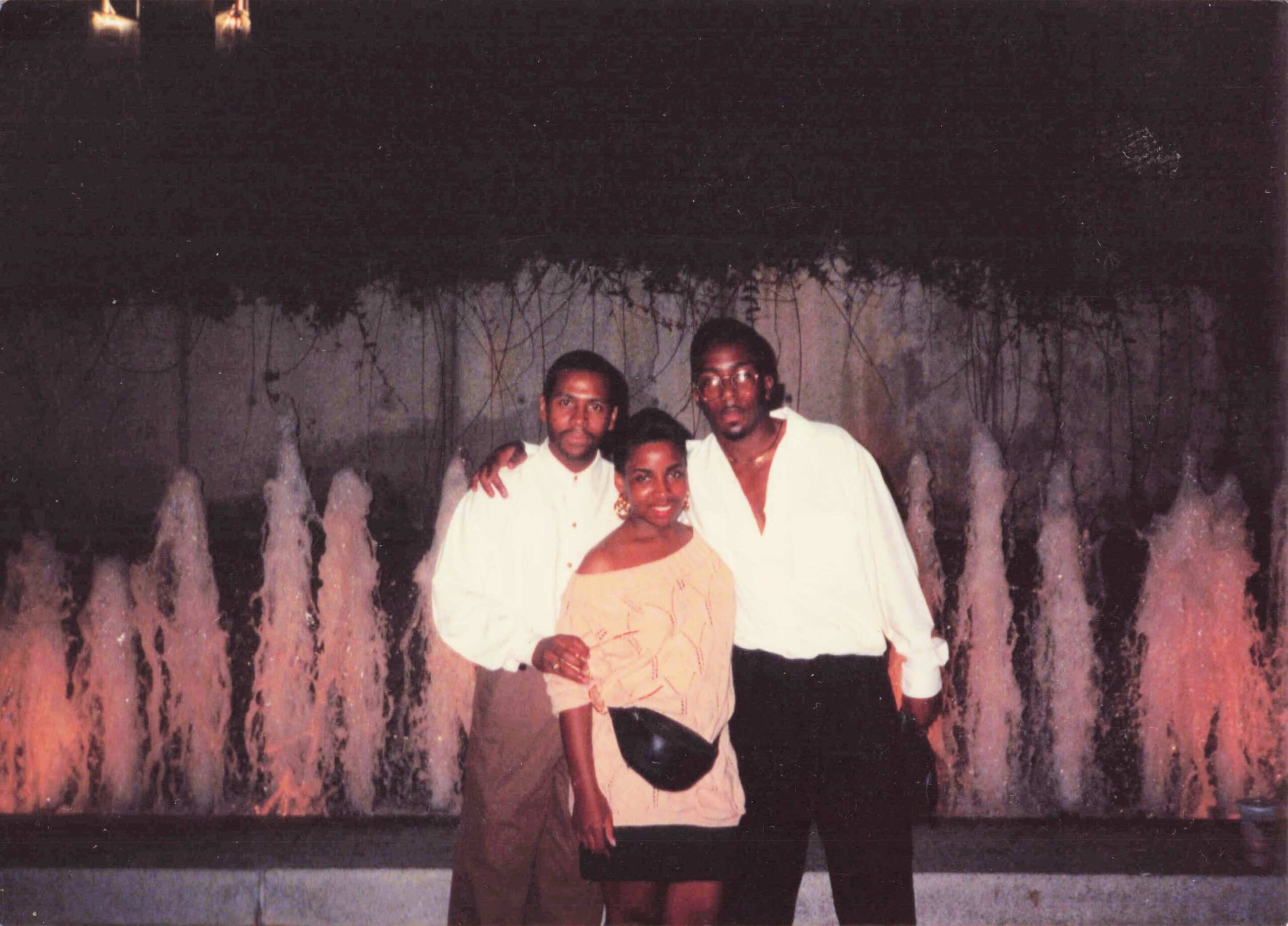 Three people pose together in front of a fountain backdrop at night; two men in white shirts and a woman in a light dress stand closely smiling.