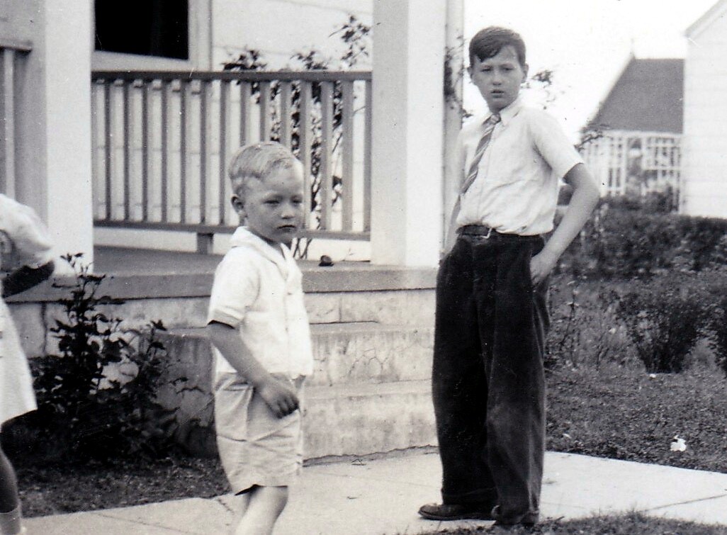 Two boys stand on a sidewalk in a suburban yard; younger boy in light shirt and shorts, older boy in a white shirt and tie watches.