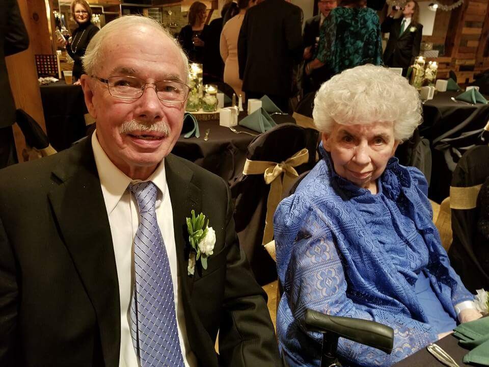 Smiling elderly man in a black suit with lavender tie sits next to an elderly woman in a blue lace dress at a formal event.