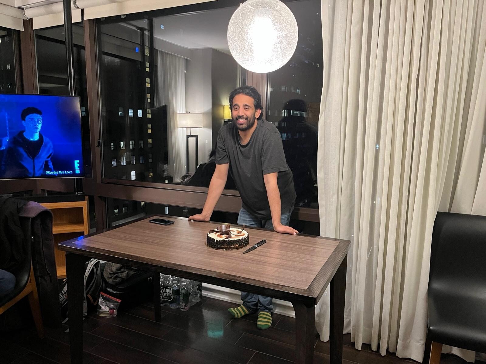 Man in a gray t-shirt leans on a dining table with a chocolate cake in front, smiling indoors at night.