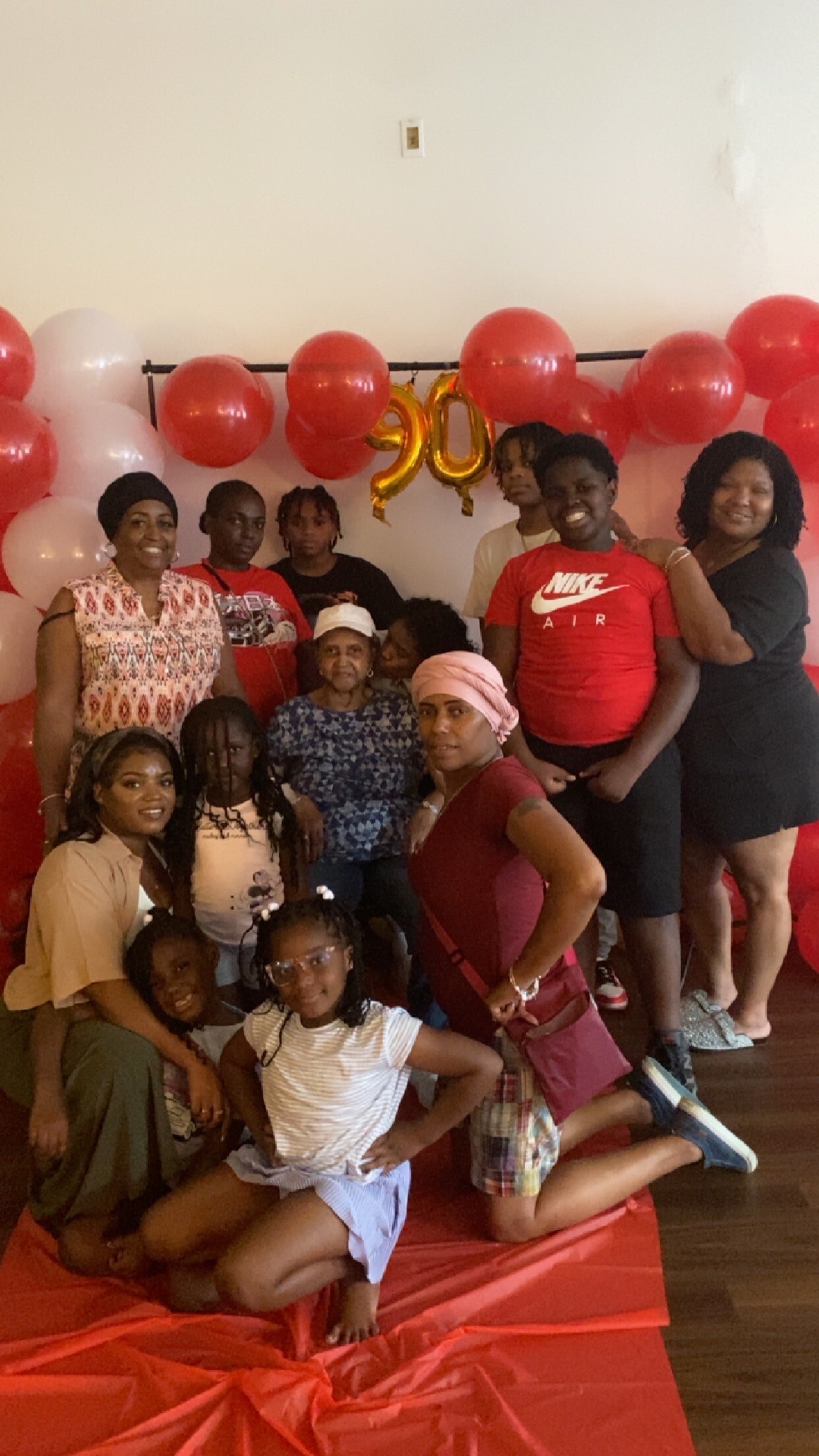 Group of family and friends posing at a celebration with red and white balloons, and gold '90' balloons in the background.