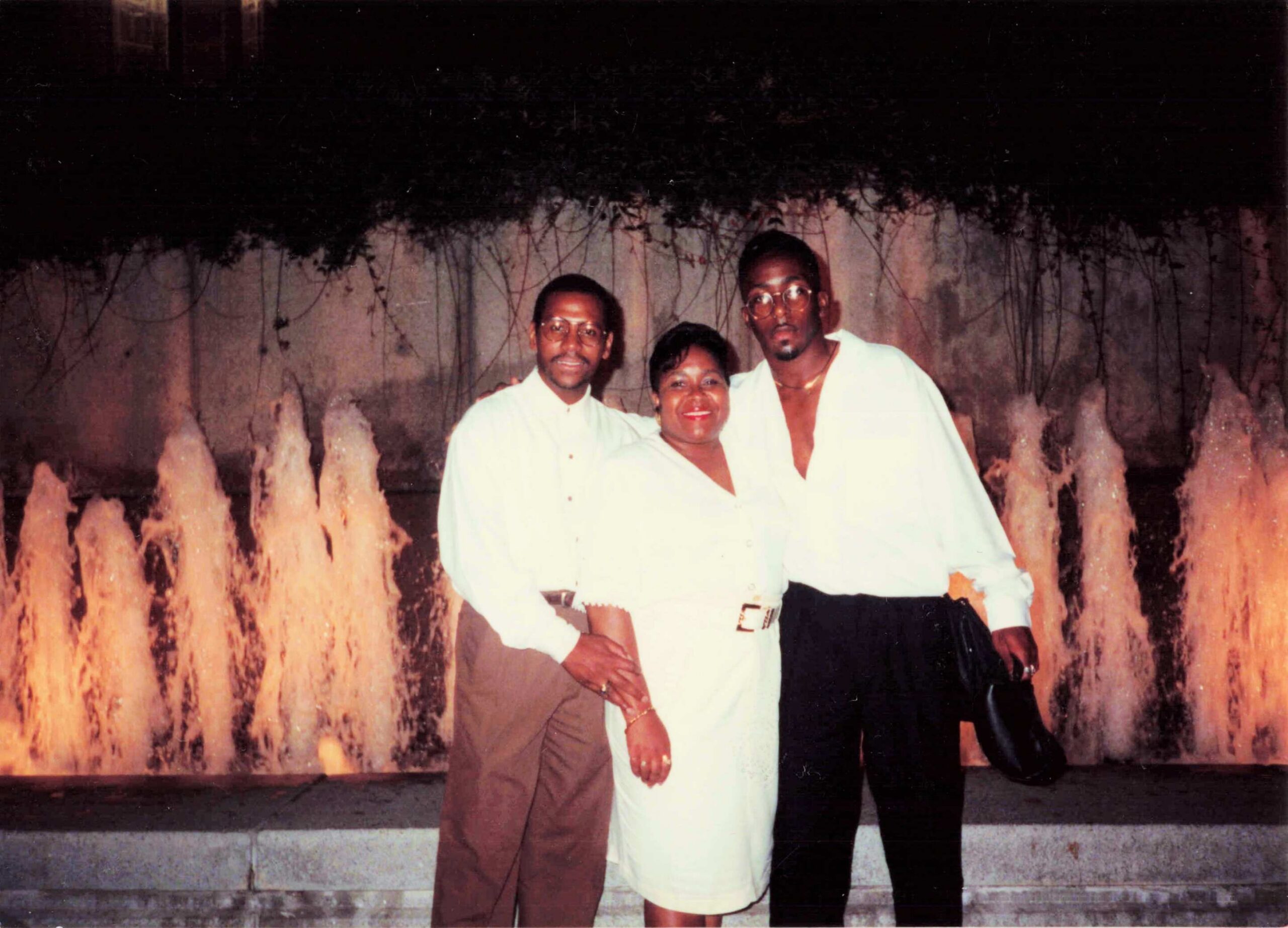 Three people posing together by a lit fountain at night, wearing white and light-colored clothing and smiling at the camera.