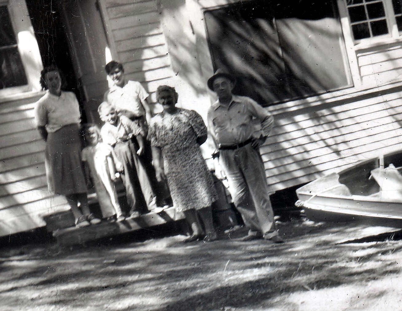 Group of adults and children posing on the front steps of a wooden house in a black-and-white photo.