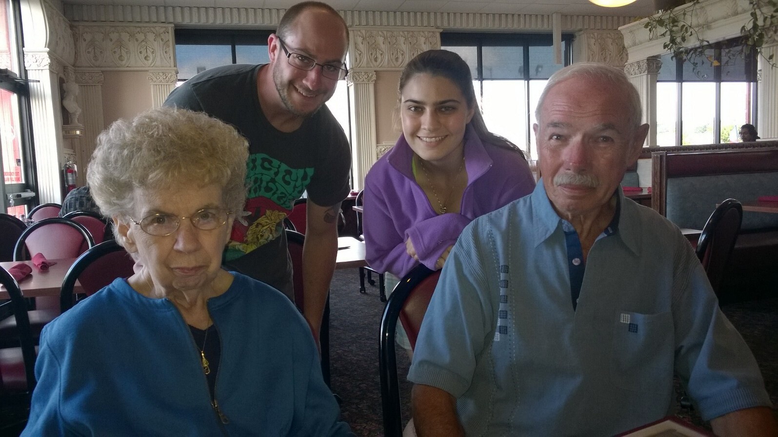 Four people posing at a restaurant table: an elderly couple in blue in the foreground, with a younger man and woman leaning in from behind and smiling in the background.