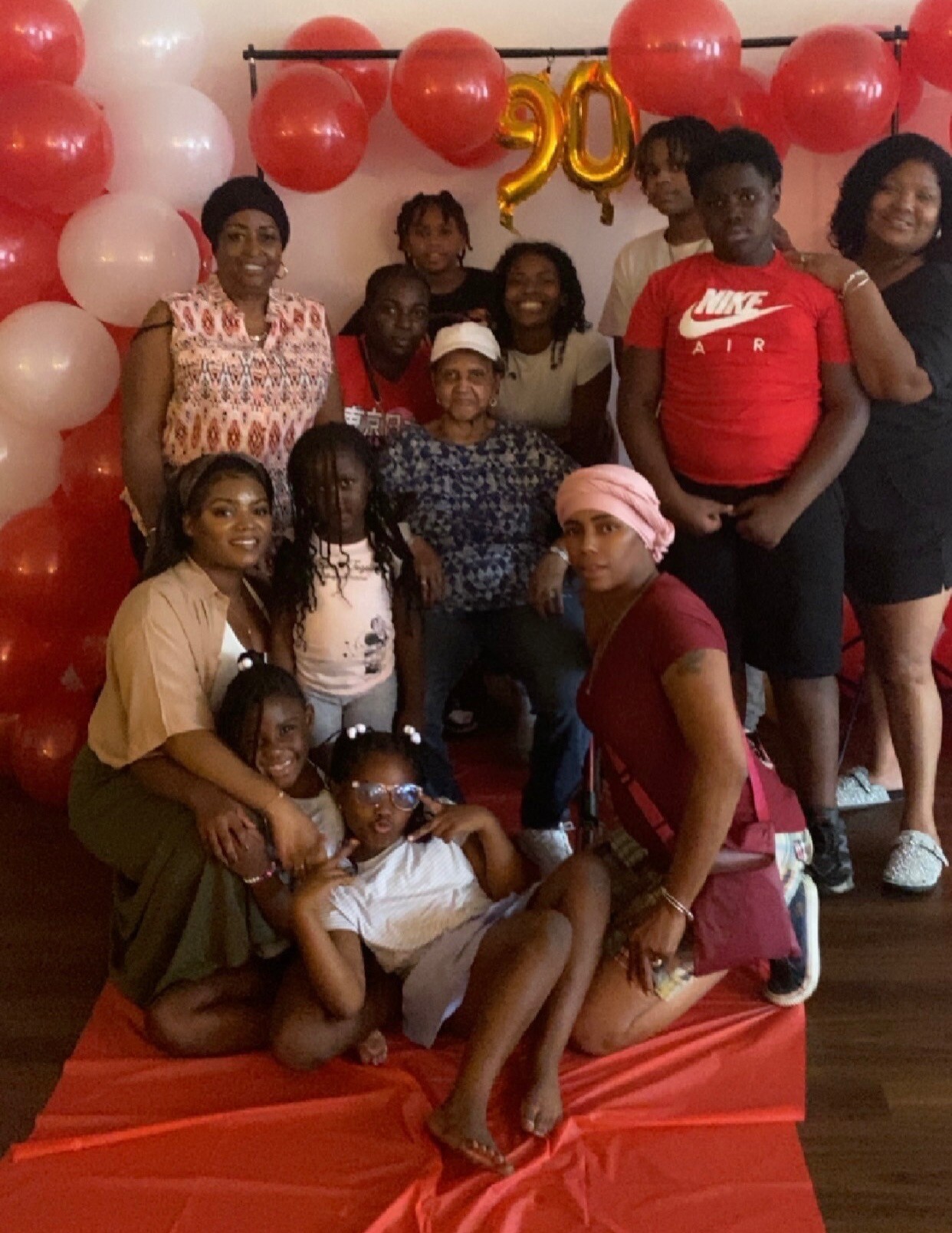 Group of friends and family posing for a birthday photo with red and white balloons and a gold '20' balloon in the background, indoors.
