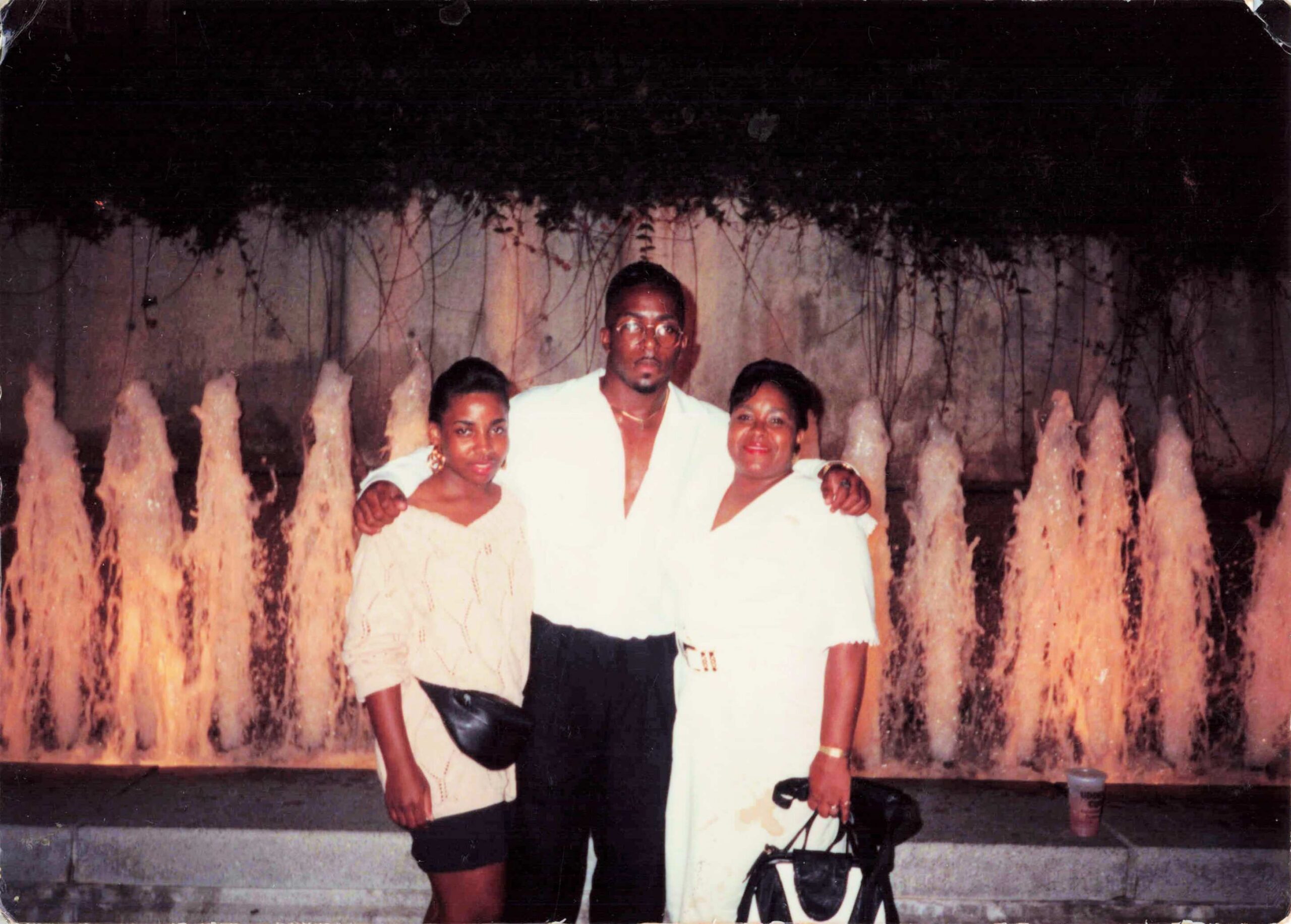 Three adults posing together in front of a vertical fountain wall, dressed in white outfits, smiling at the camera.