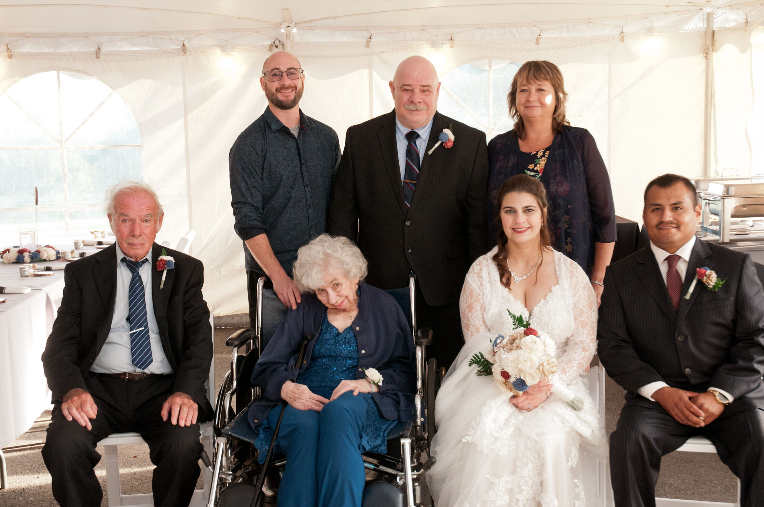 Bride in a lace wedding dress holding a bouquet sits at the front, flanked by wedding guests under a white tent.