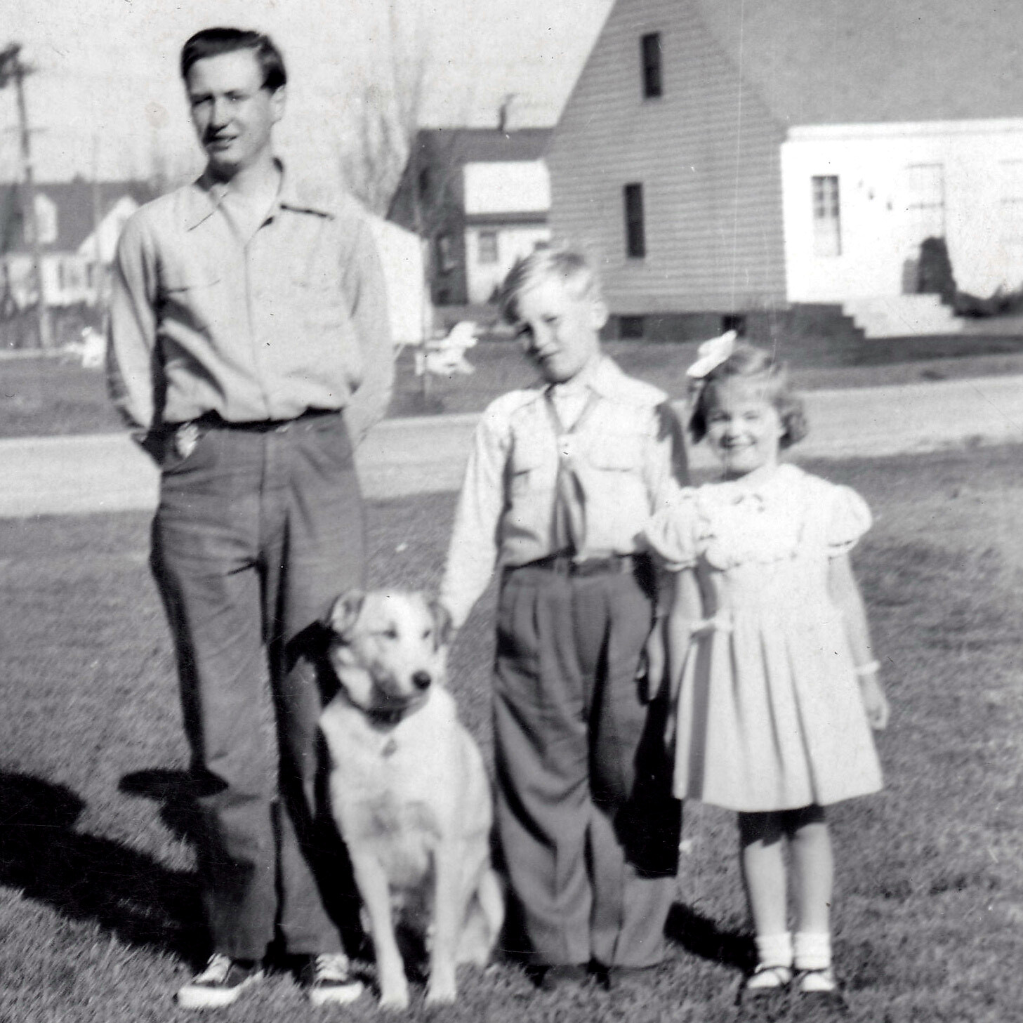 A man, two children, and a dog stand on a lawn in front of suburban houses, posing for a photo.
