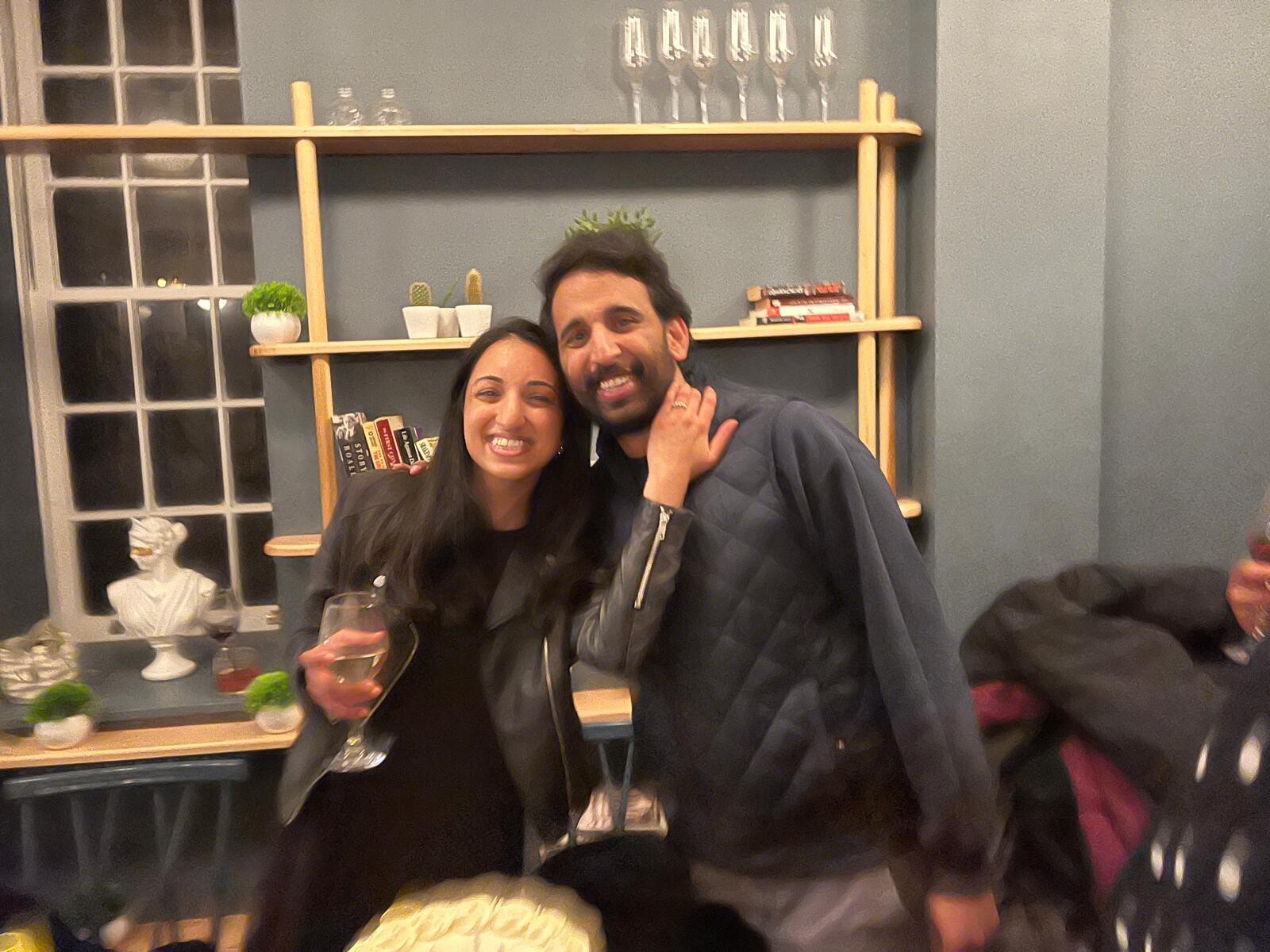Two friends posing together and smiling at a party, holding wine glasses, with a shelf and plants in the background.