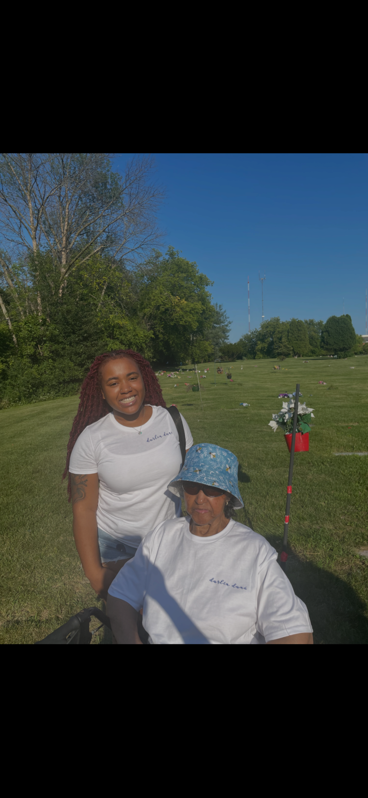 Two women pose together in a sunny cemetery-like park; one in a wheelchair wearing a blue hat, the other standing in a white T-shirt, both smiling amid green grass and scattered flowers or tributes.