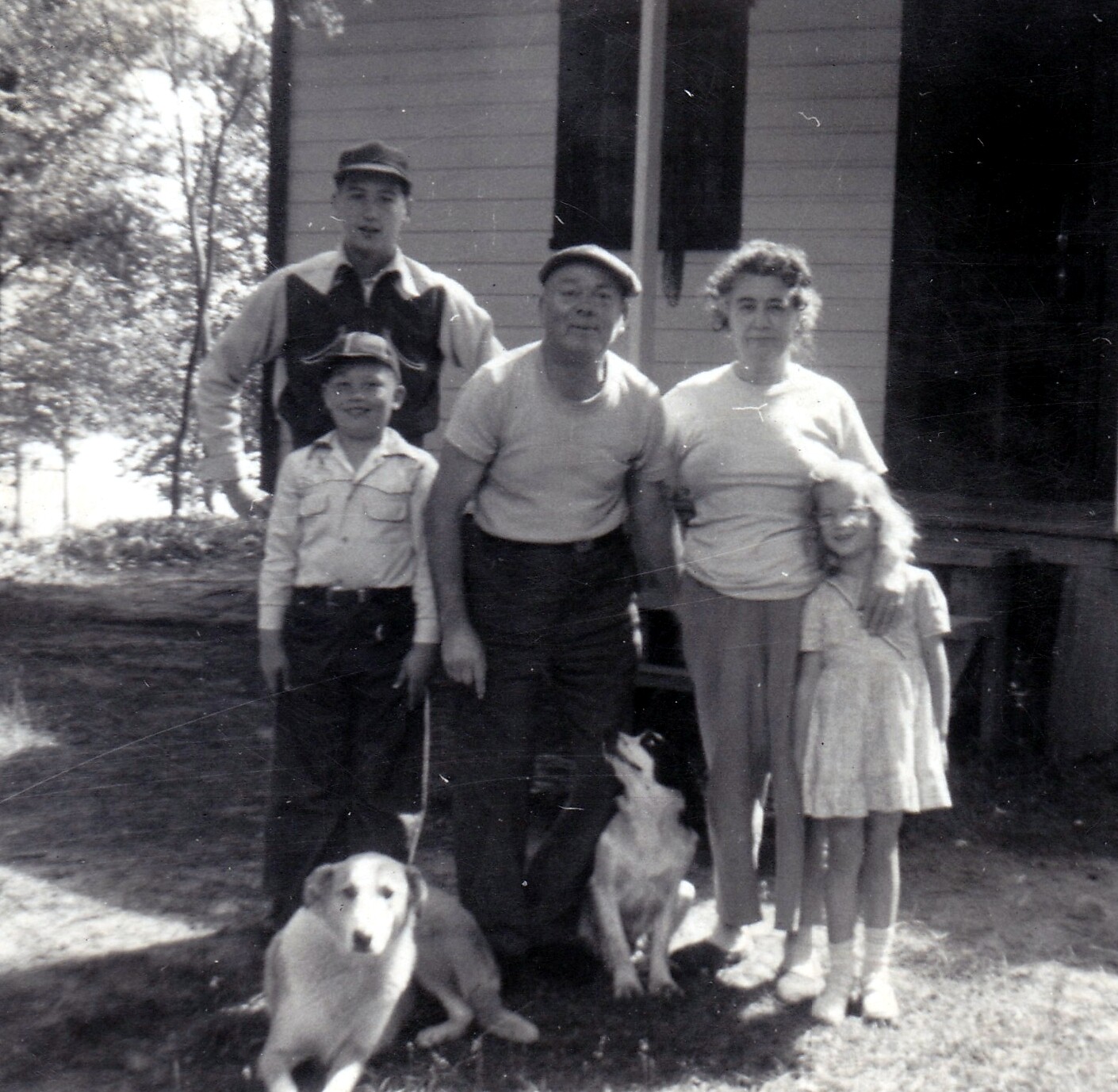 Family of five posing outdoors in front of a house, with two dogs at their feet.