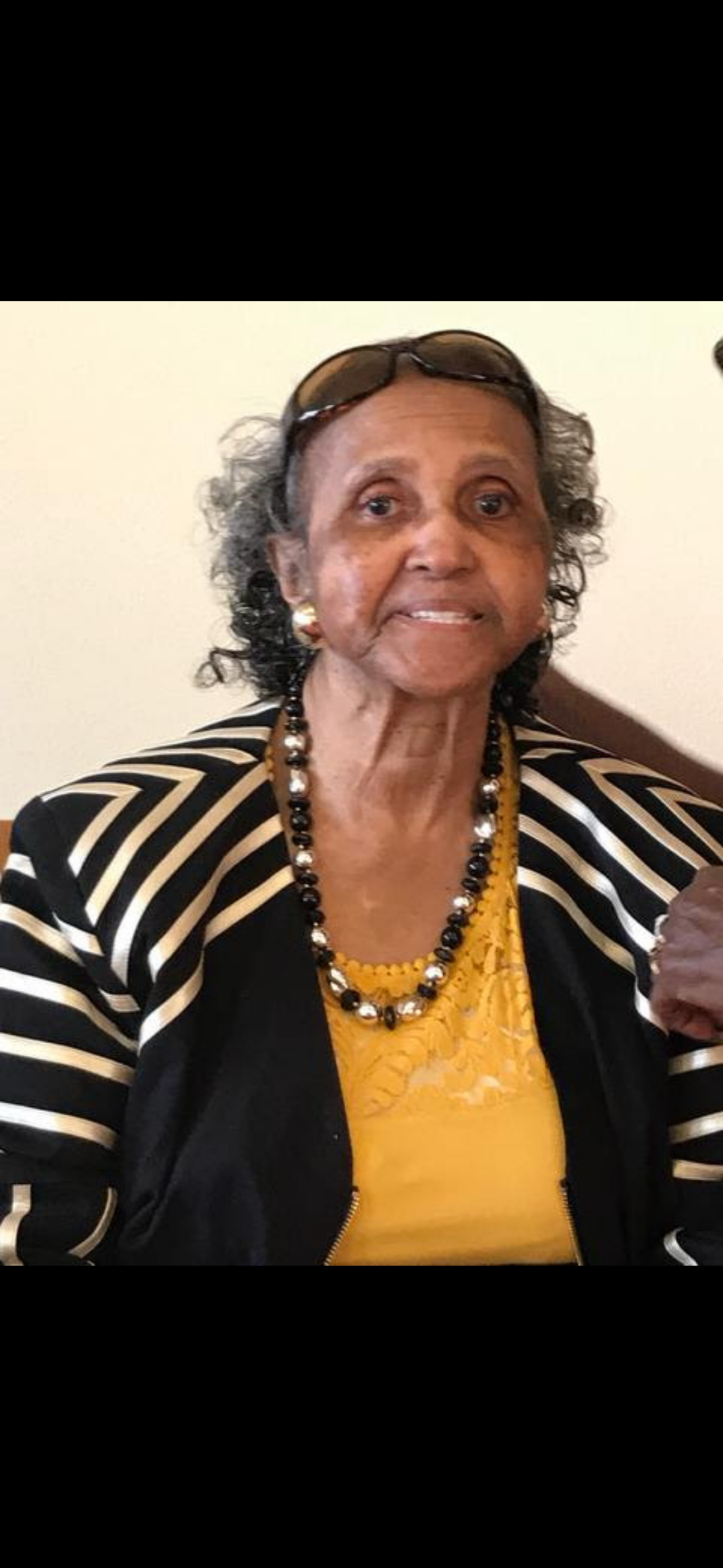 Portrait of an elderly woman with short gray hair, glasses on her head, wearing a yellow top and a black-and-white striped jacket, with a beaded necklace.