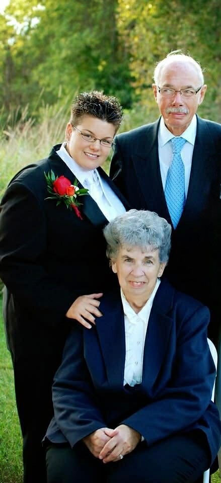 Three-generation family portrait outdoors: seated elderly woman in a navy blazer, with a middle-aged woman in a black suit and a red boutonniere standing beside her and an older man in a suit and blue tie behind them.