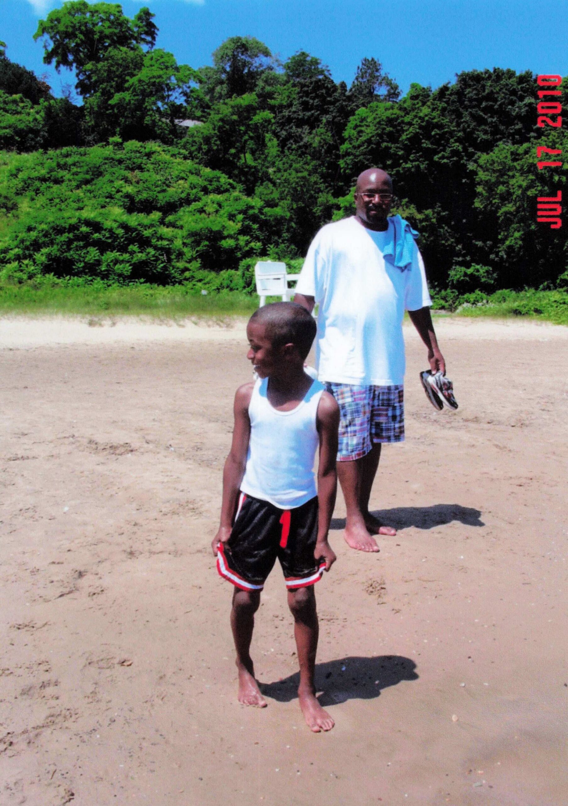 Man and boy walking on a sandy beach with green bushes behind them; the man holds flip-flops while the boy walks ahead.