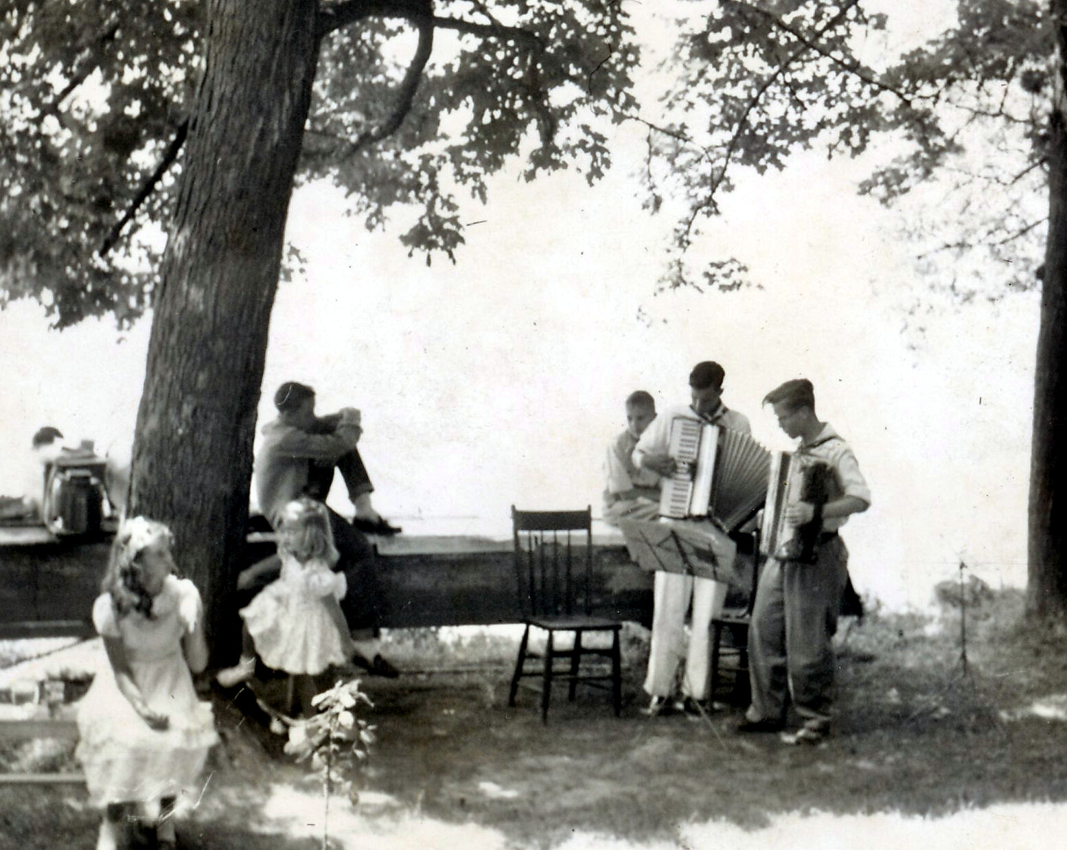 Black-and-white photo of a lakeside gathering: two men play accordions while children in dresses watch under a tree by a bench and wall.