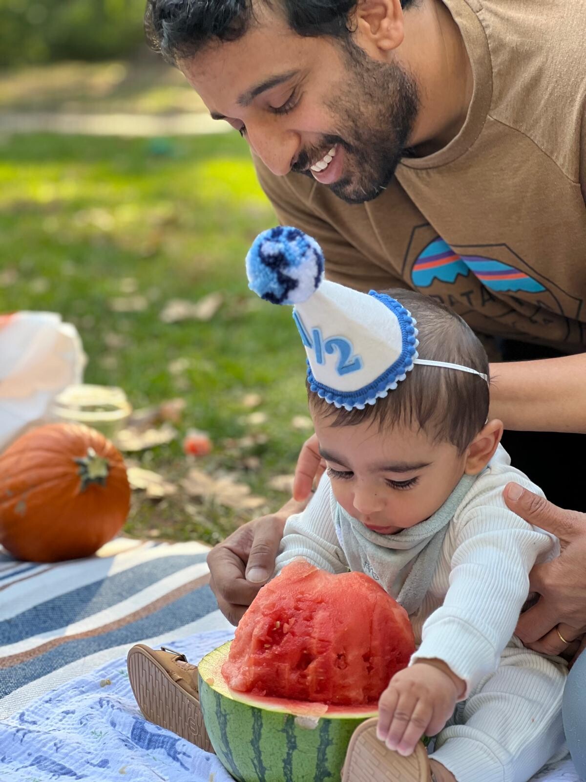 Father leaning over a baby wearing a blue and white birthday hat, feeding watermelon at a park picnic.