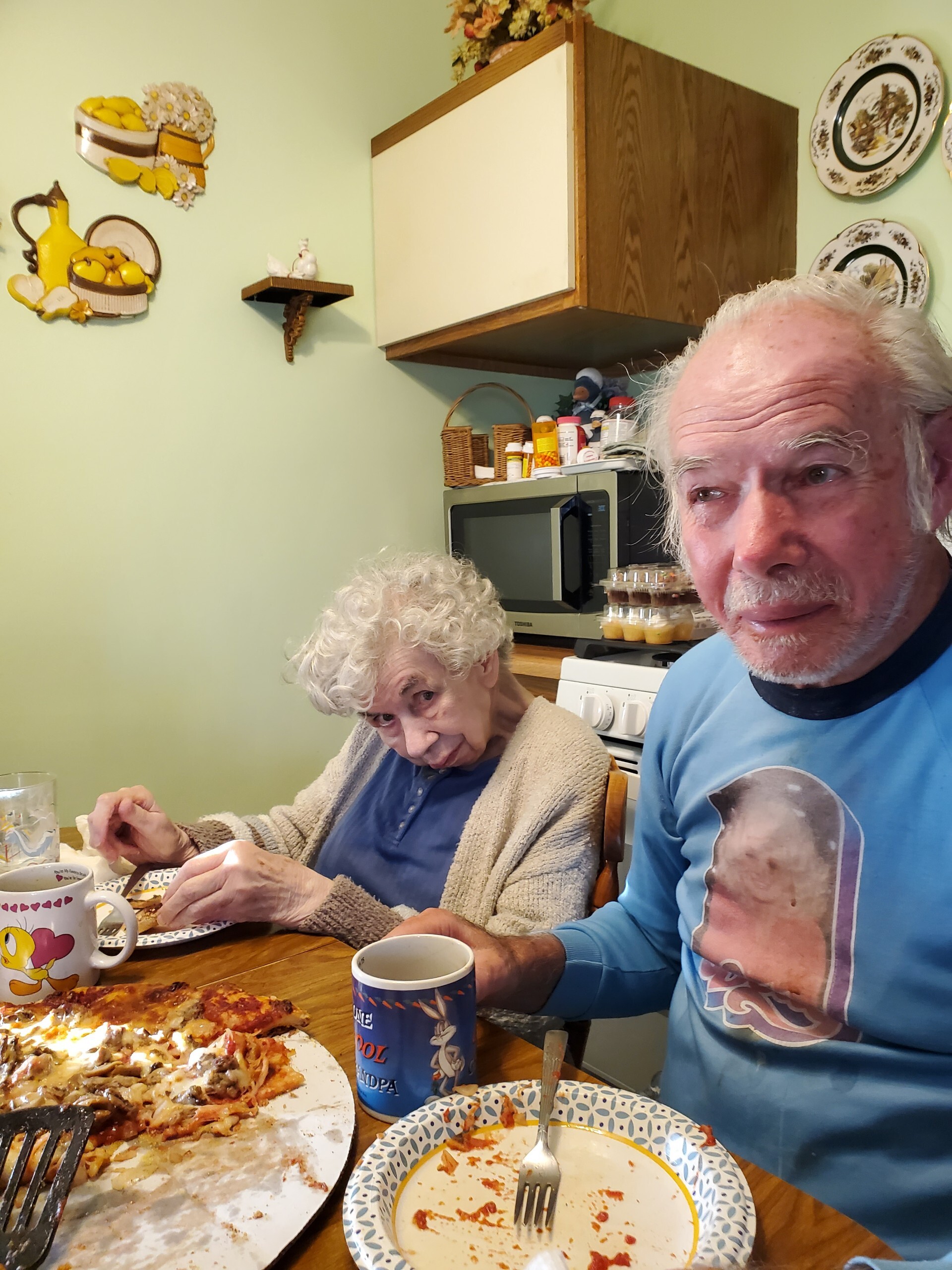 Two elderly people sit at a kitchen table eating pizza, with plates and mugs nearby in a cozy home setting.