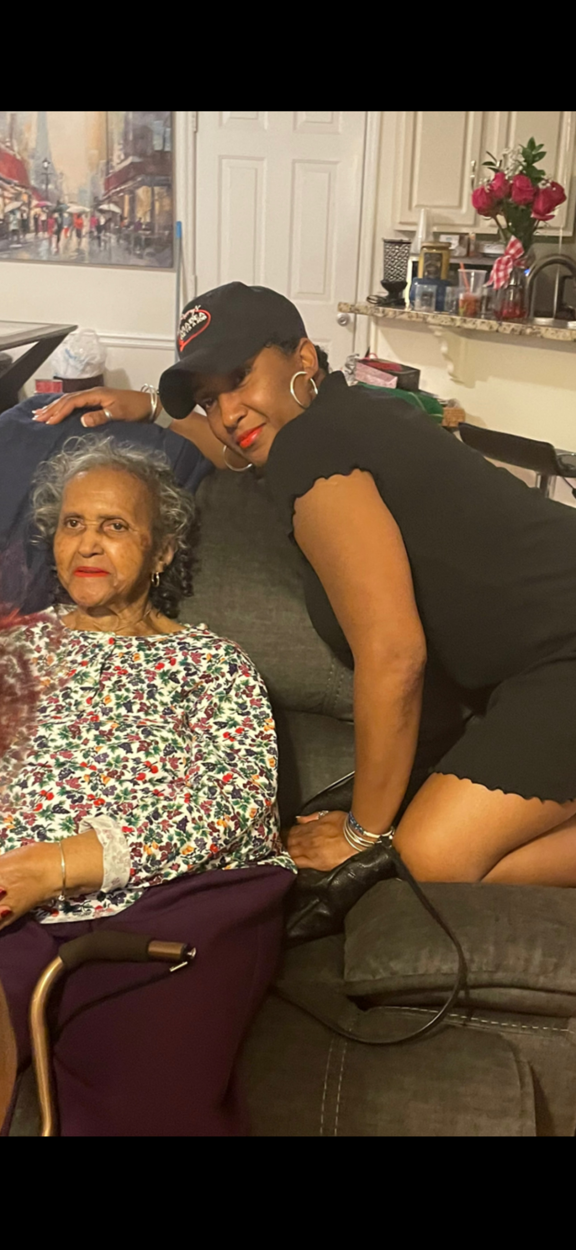 Woman in a black outfit and cap leaning over a senior woman seated on a sofa in a living room, both smiling.