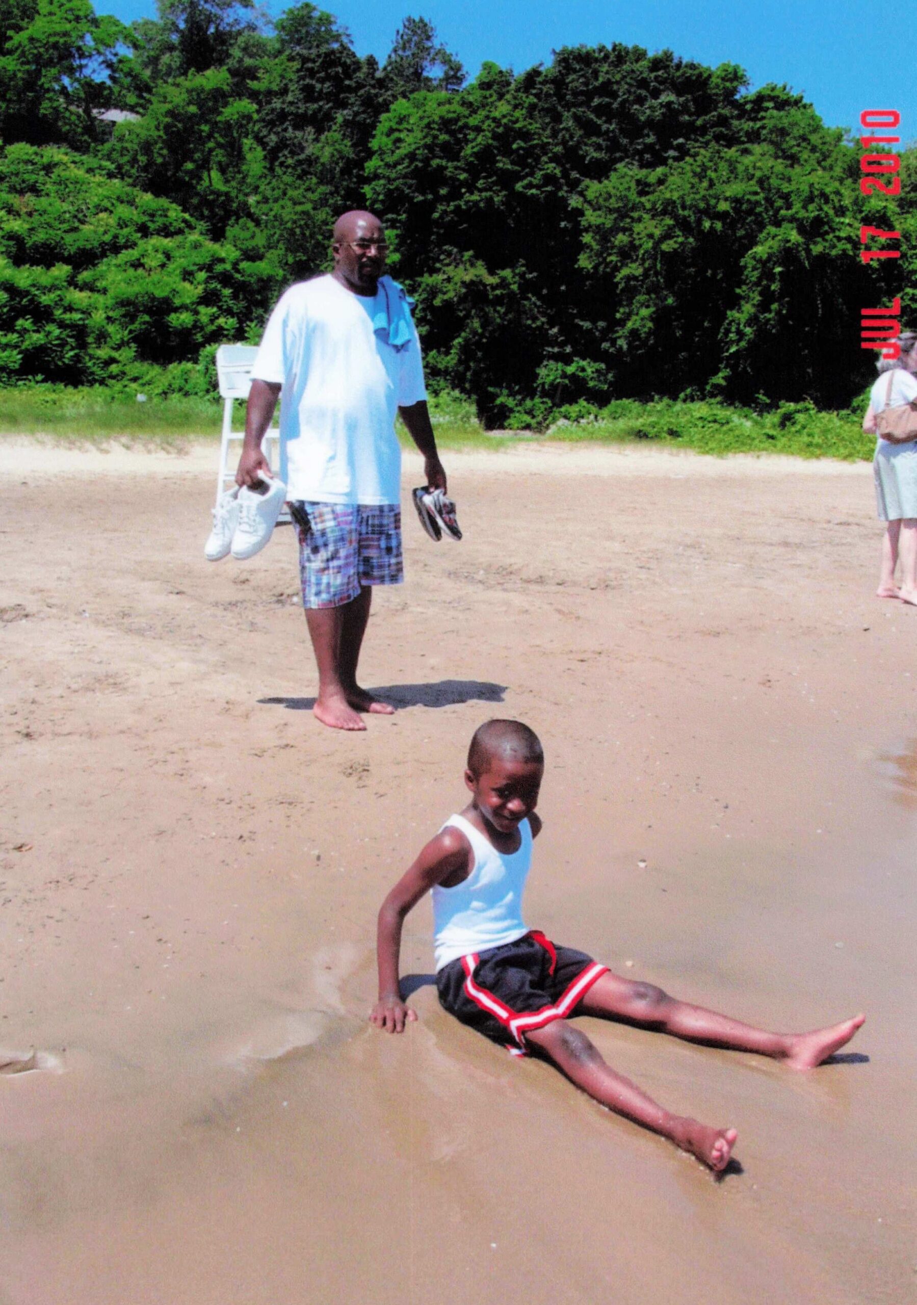Man on a sunny beach holding a pair of shoes and a child sliding in the sand nearby, with trees in the background.