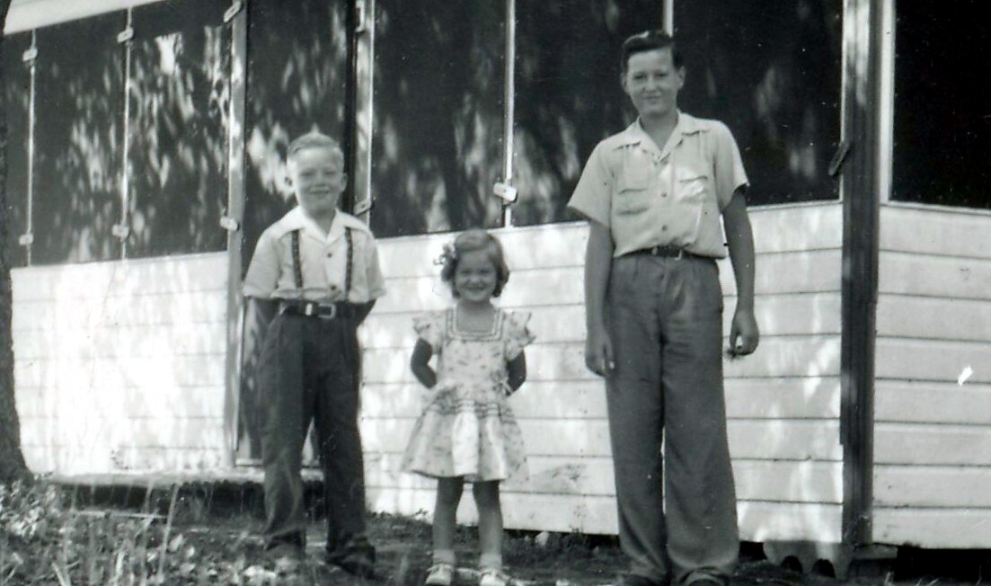 Three children posing outdoors in front of a white wooden building; a boy on the left, a girl in the middle, and a woman on the right.