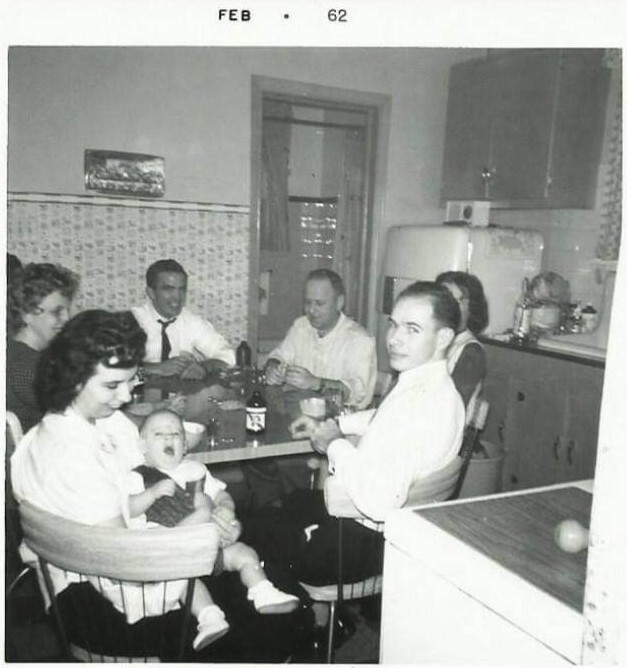 Black-and-white family gathering around a dining table in a mid-century kitchen, a baby resting on a woman's lap.