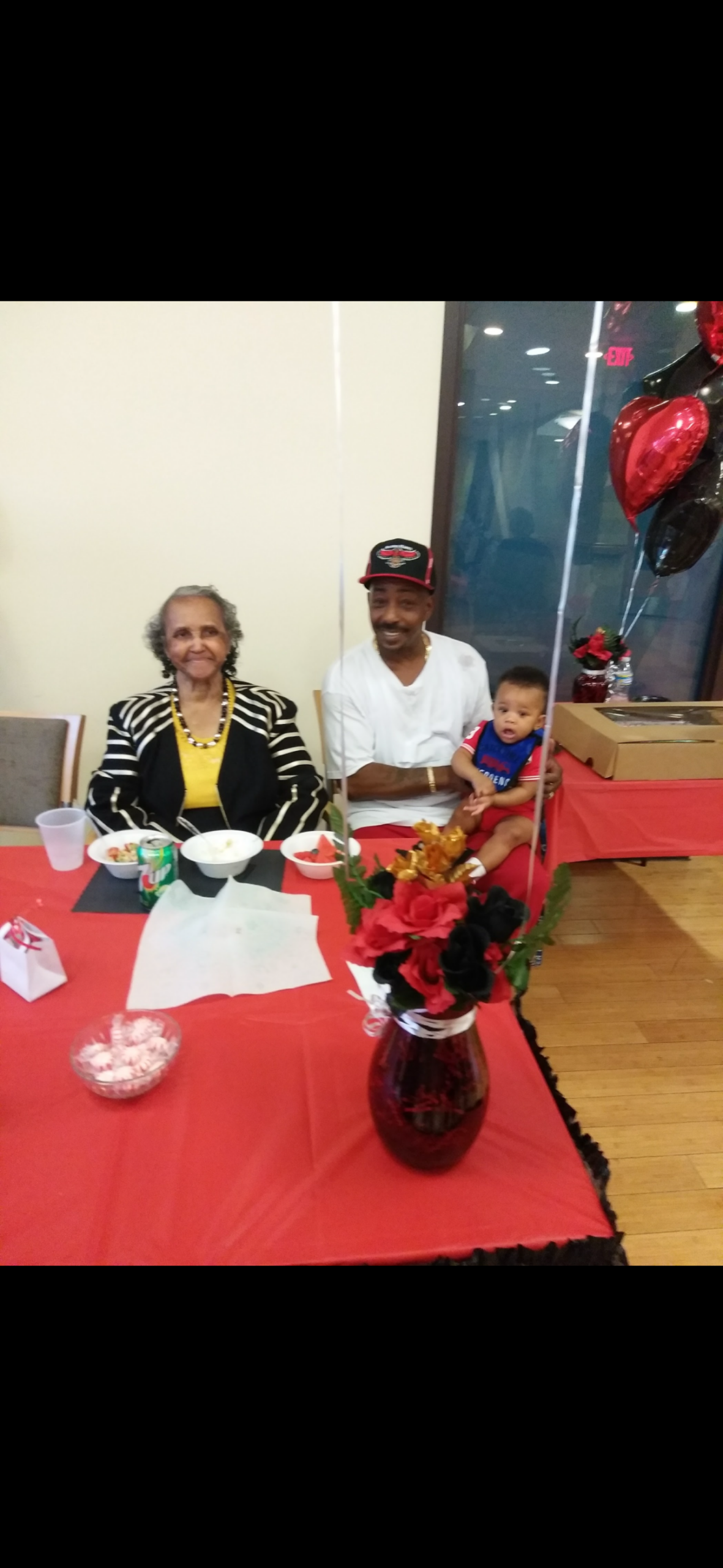 Smiling grandmother, father, and toddler sit at a red party table with balloons and a flower centerpiece.