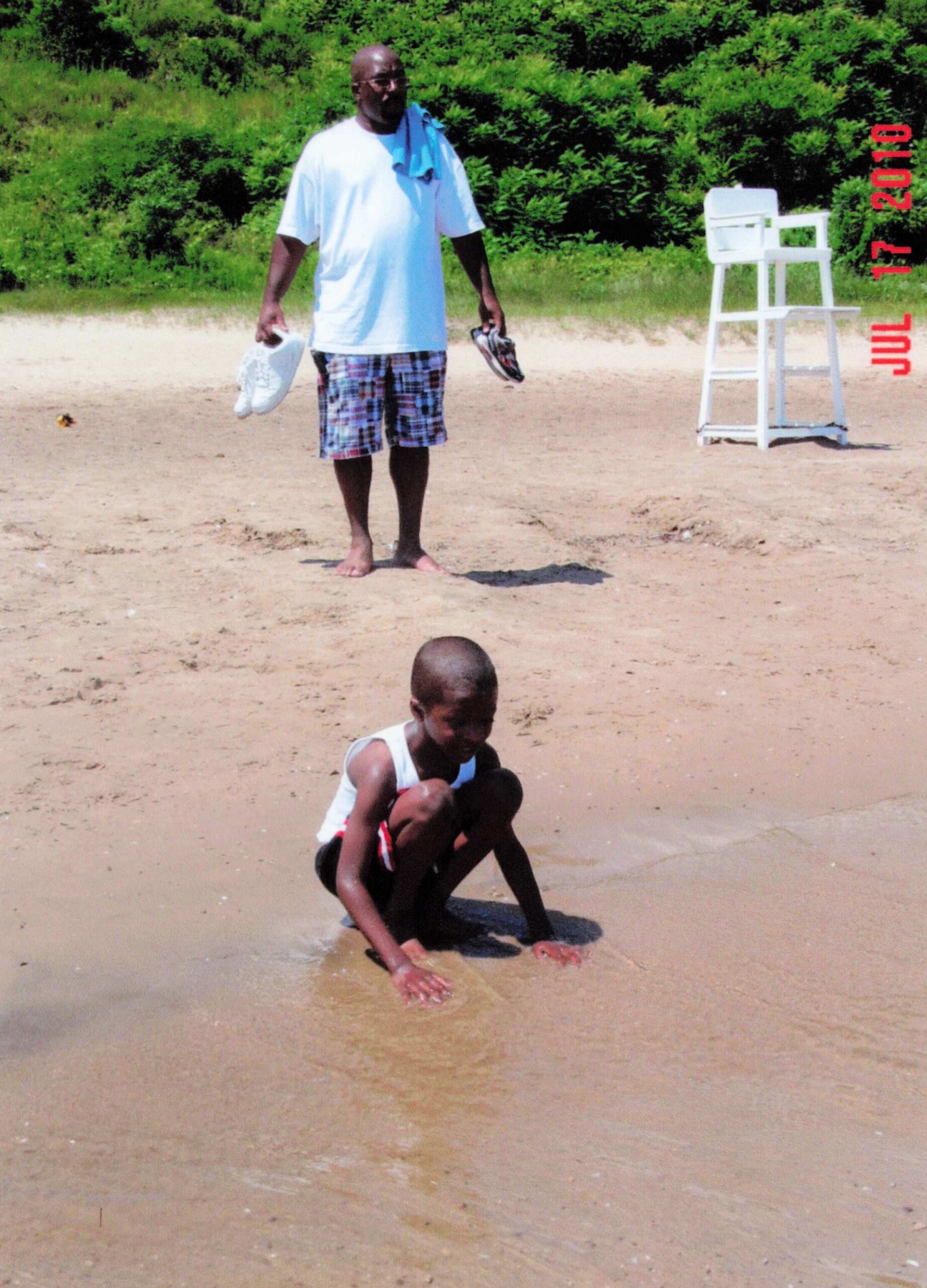 Man in a white shirt and plaid shorts stands barefoot on a sandy beach, holding shoes; a boy plays in shallow water nearby with a white lifeguard chair to the right.