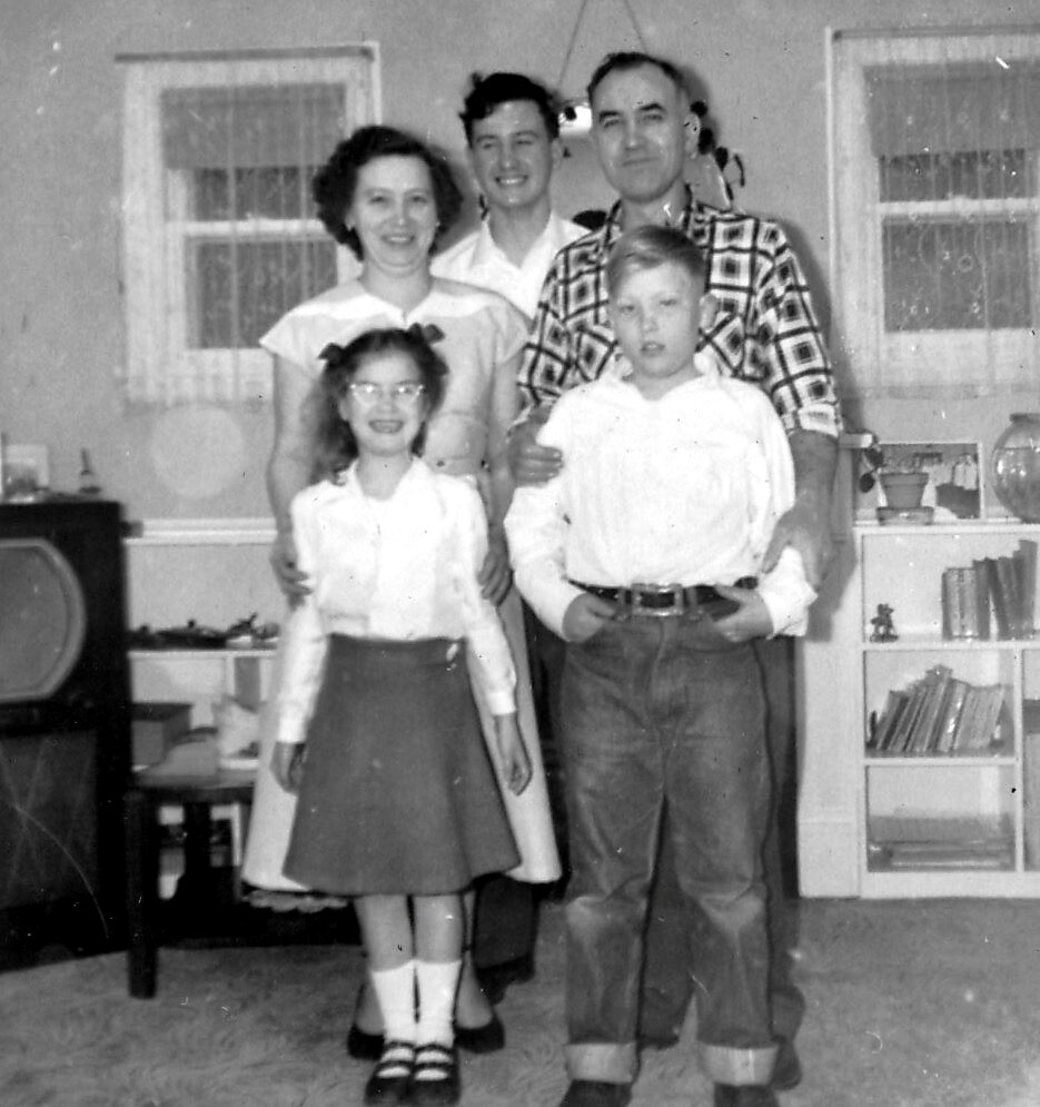 Black-and-white family photo in a living room: two adults stand behind with two children in front—a girl with glasses and a boy in a white shirt—posing for the picture.