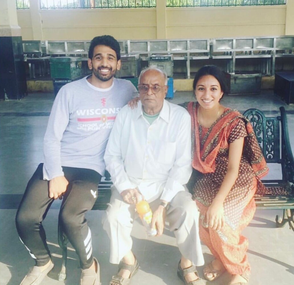 Three people sit on a bench together: a smiling young man on the left, an elderly man in the middle, and a smiling woman in traditional dress on the right.