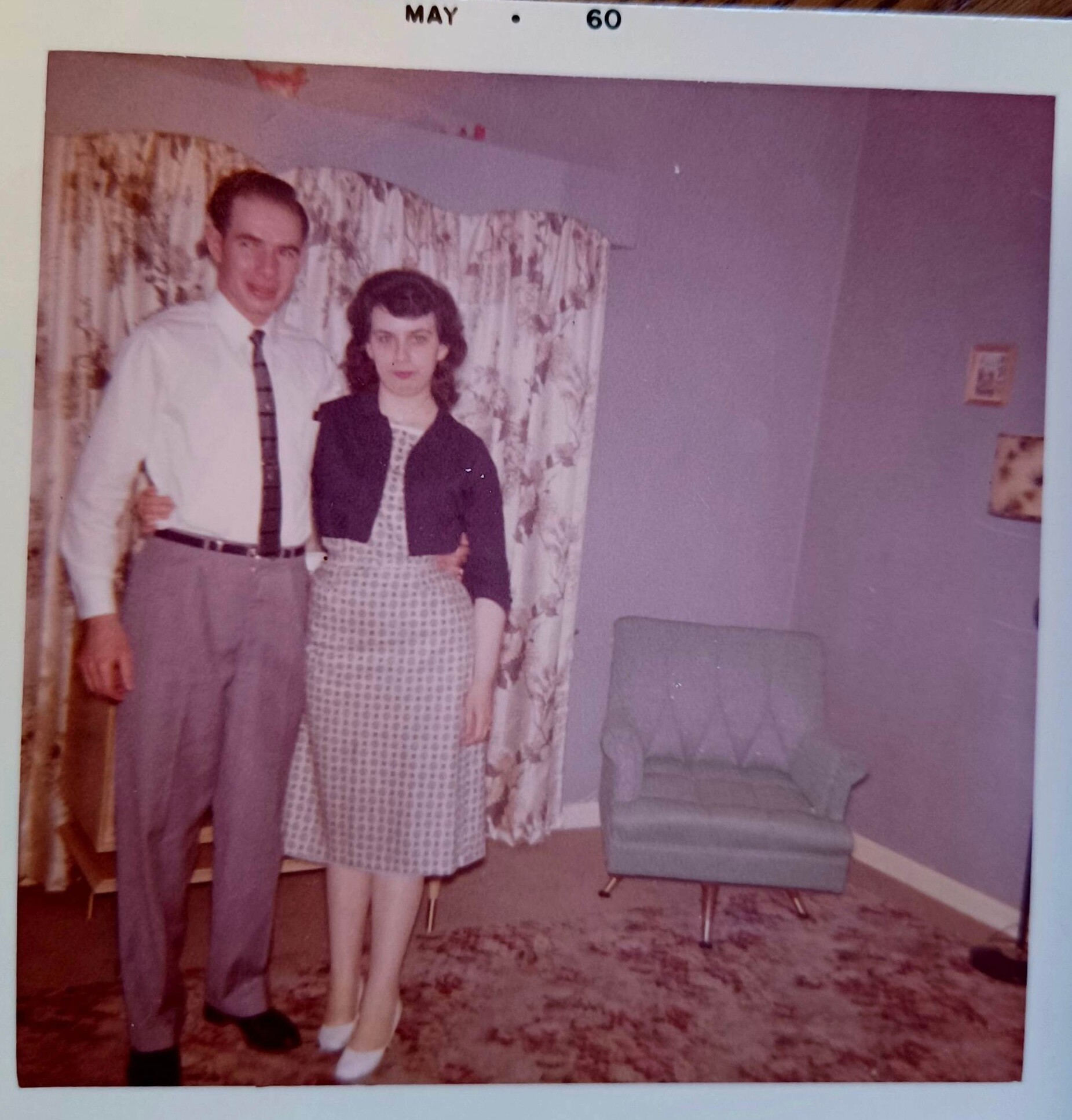 Man in a white shirt and tie stands beside a woman in a checkered dress in a 1960s living room; May 60 date stamp visible at top.